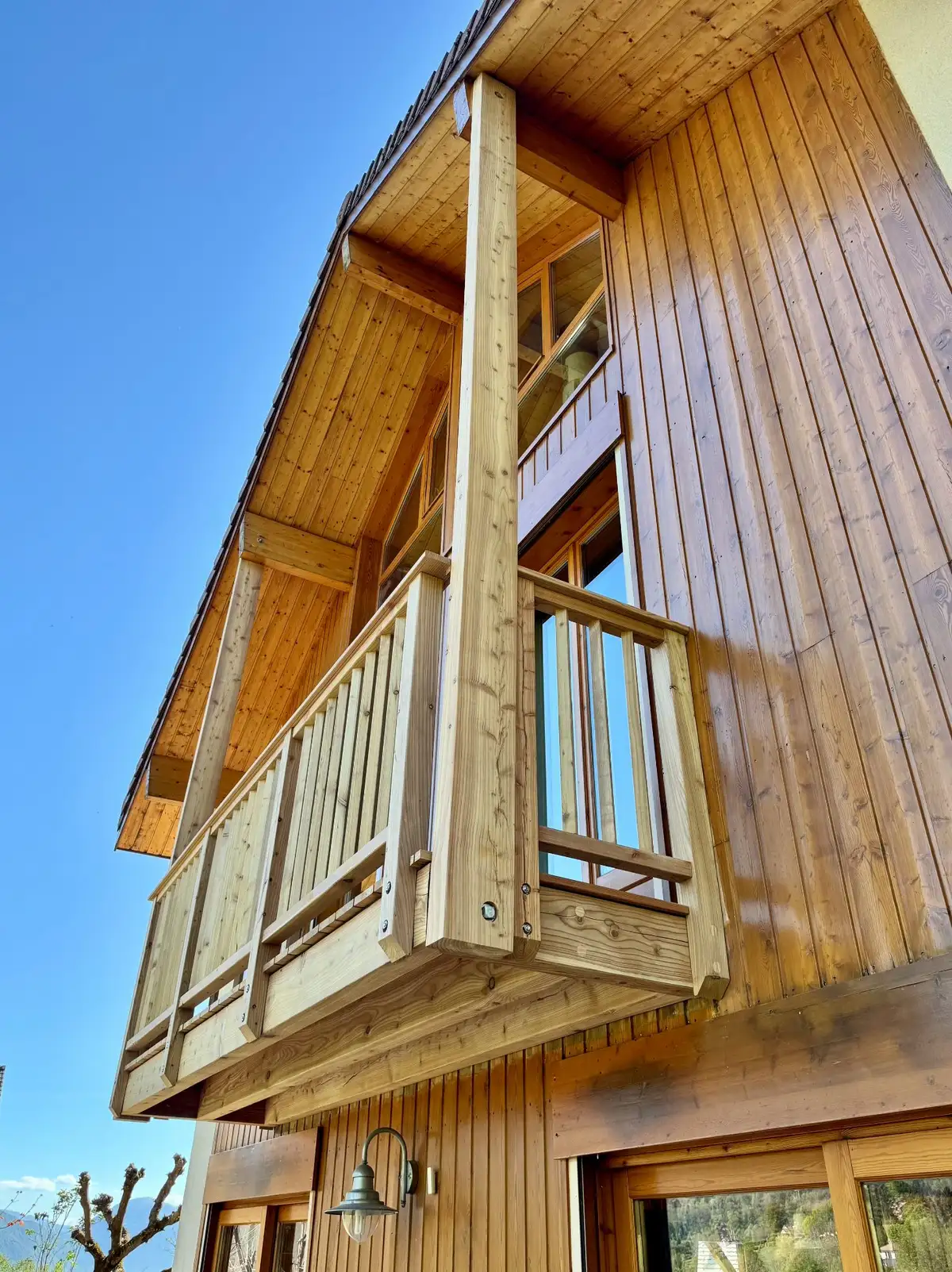 Balcon en bois suspendu à la façade d'une maison en bardage bois vertical, vu en contre-plongée sur ciel bleu.