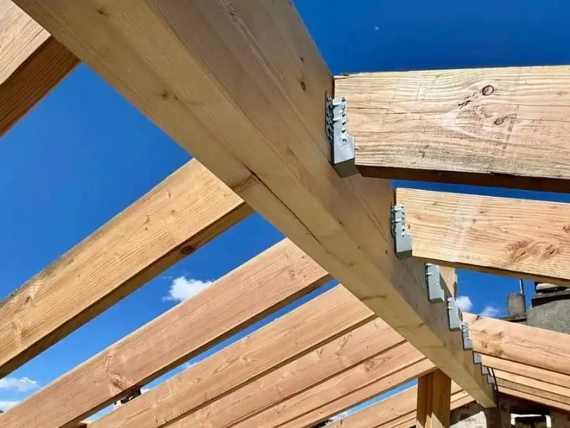 Charpente neuve en bois clair vue de l'intérieur du comble, pannes et chevrons assemblés sur ferrures se découpant sur un ciel bleu.