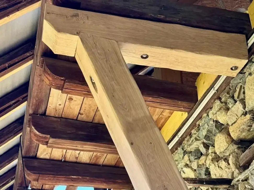 Vue en contre-plongée sous l'avancée d'un toit : une console de chêne clair fraîchement taillée soutient les chevrons anciens en bois sombre, contre un mur de pierres apparentes.