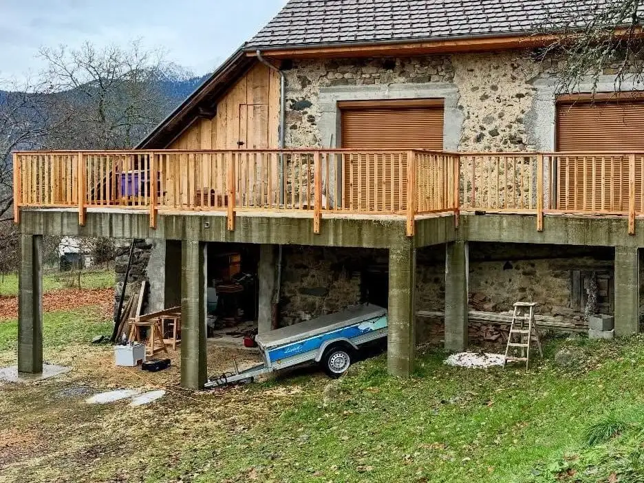 Grange de pierre à flanc de pré, dont la terrasse sur pilotis de béton est bordée d’un garde-corps neuf en bois clair à barreaux verticaux ; montagne boisée à l’arrière-plan.