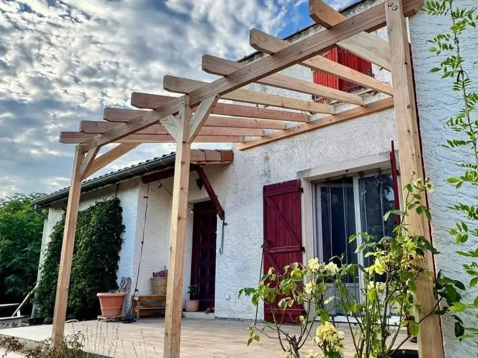 Pergola en bois clair adossée à une maison blanche à volets rouges, ses chevrons se détachant sur un ciel nuageux ; rosier grimpant au premier plan.
