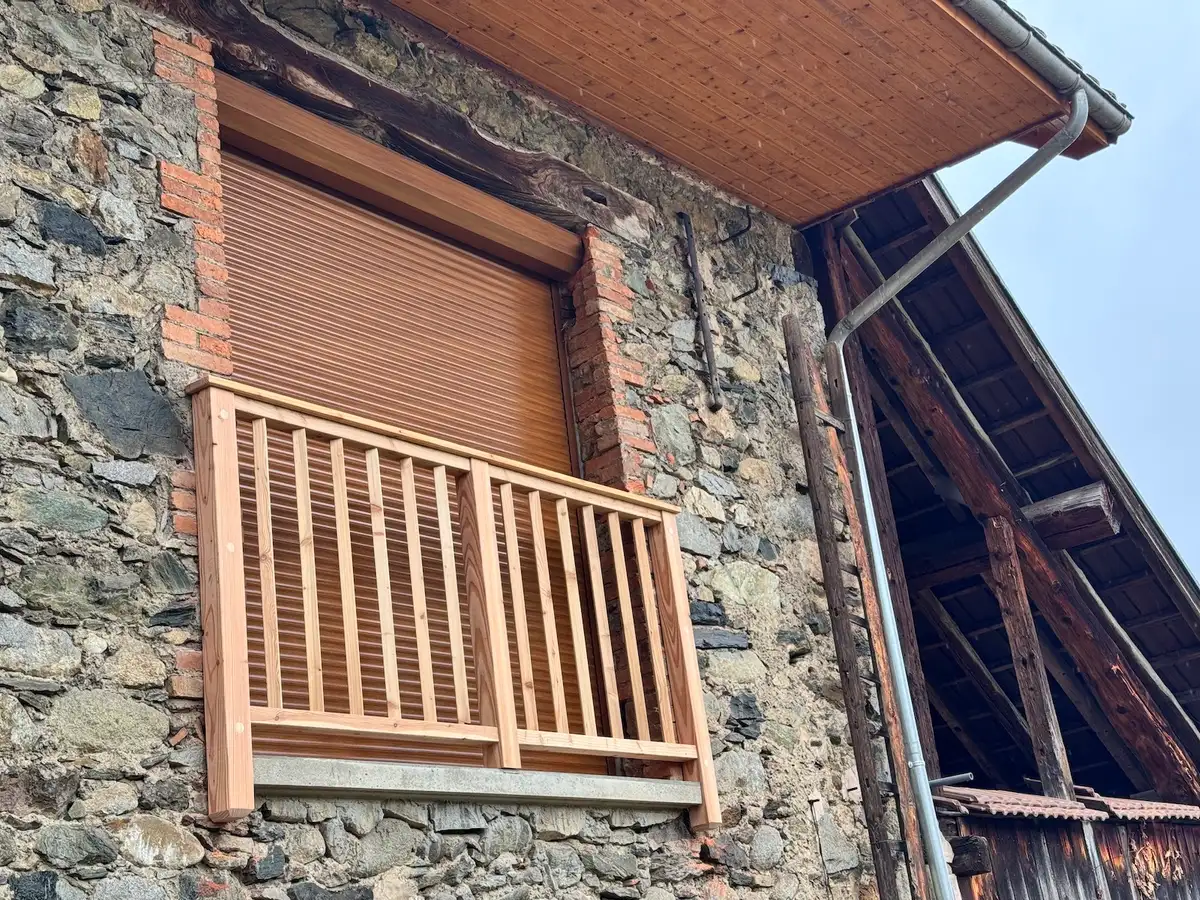 Grange de pierre à flanc de pré, dont la terrasse sur pilotis de béton est bordée d'un garde-corps neuf en bois clair à barreaux verticaux ; montagne boisée à l'arrière-plan.